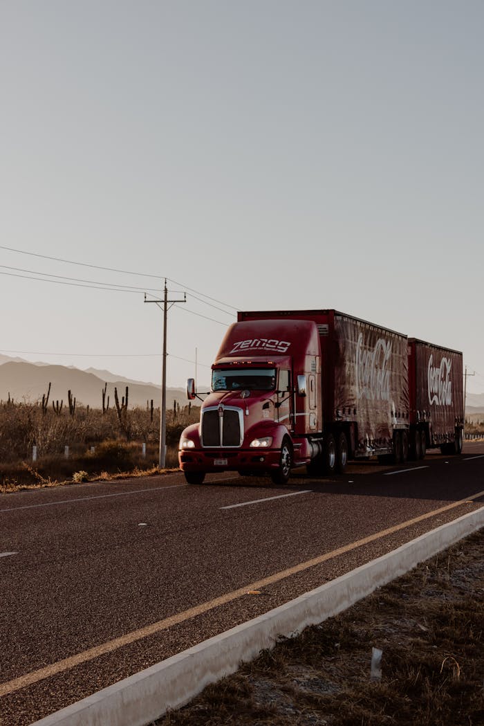 who-we-are A large red truck transports goods along a desert highway under a clear sky.