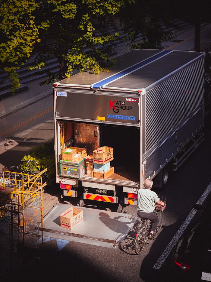 journey A delivery truck unloading boxes in a city street, with a bicyclist passing by on a sunny day.